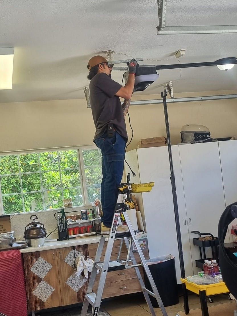 A technician on a step ladder working on a garage door opener mounted to the ceiling, with the opener rail running back across the garage