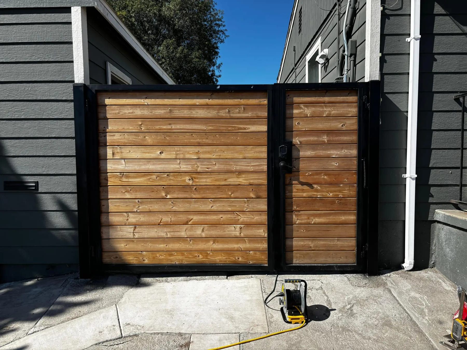 A black-framed double driveway gate with horizontal wood slats and a keypad entry panel, installed between two houses