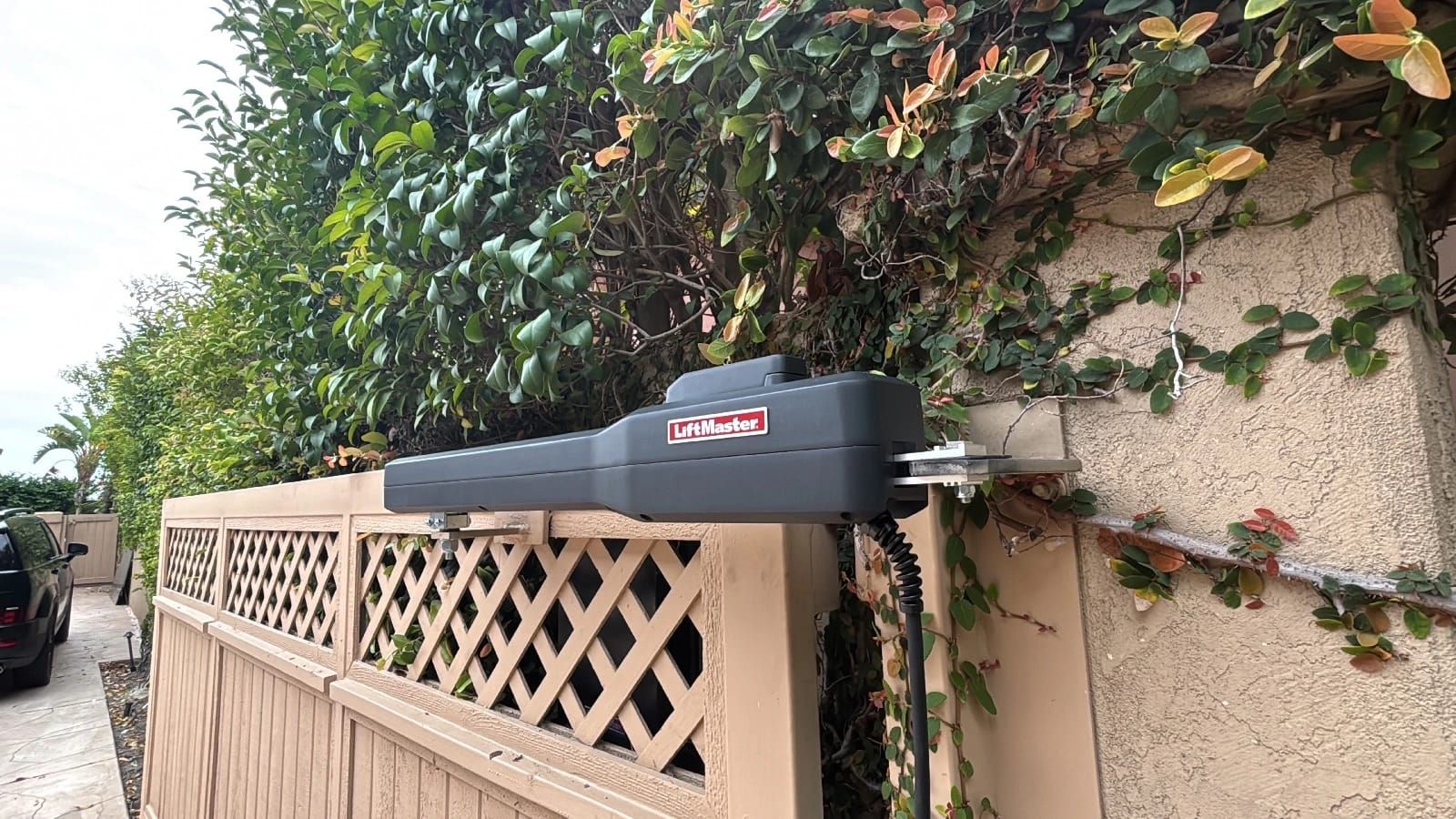 A black LiftMaster swing gate operator mounted on a stucco wall above a lattice topped fence, its arm extending over the gate