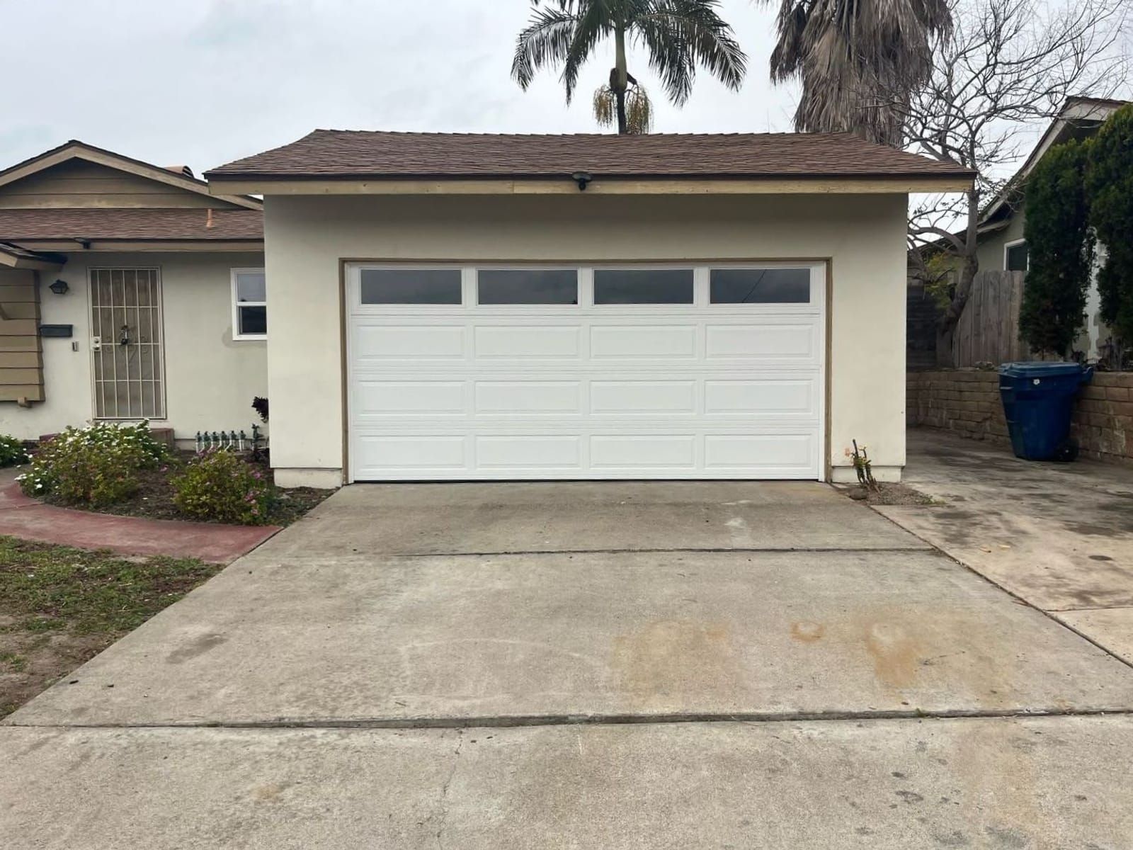 A newly installed white sectional garage door with a row of four window panes, on a detached garage with cream stucco walls and a shingle roof