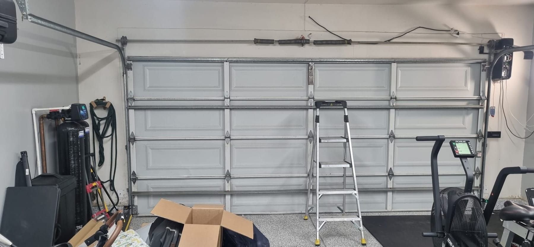 A closed white sectional garage door seen from inside, with the torsion bar and springs mounted across the header above it and a step ladder standing in front